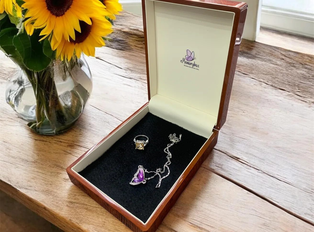 Open walnut eMemorybox on a wooden table with a ring and necklace inside, sunflowers in a vase